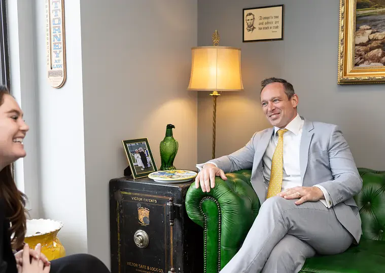 Two people in a professional office, one seated on a green leather couch, the other smiling, with a safe and lamp in the background.
