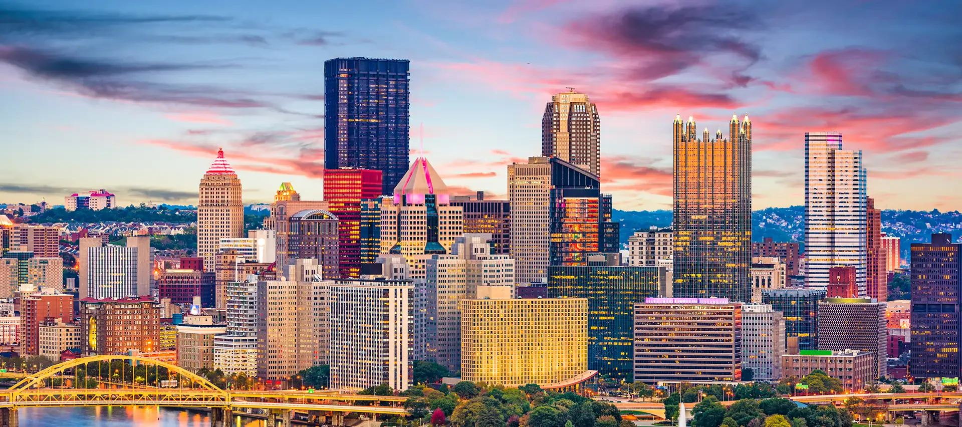 Colorful sunset over Pittsburgh skyline with the yellow Roberto Clemente Bridge in the foreground and vibrant city buildings.