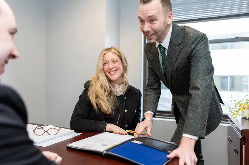 Three business professionals discussing documents in an office with a window and blinds in the background.