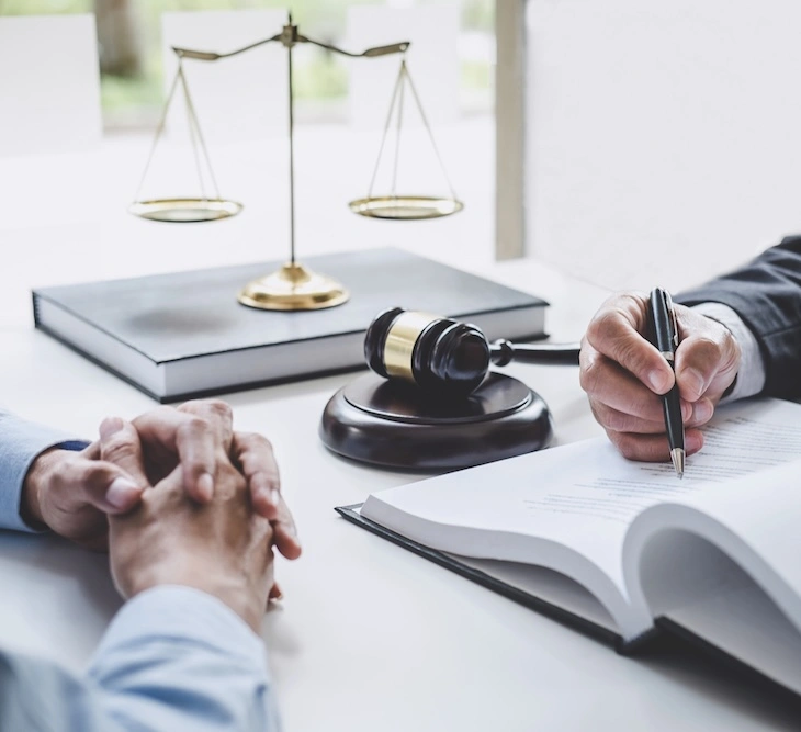 Two people sit at a desk with legal documents, a gavel, and scales of justice, while one person writes on a clipboard.
